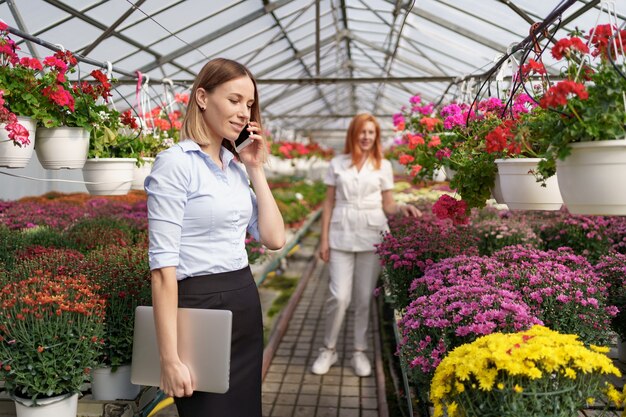 Empresária discutindo ao telefone uma proposta. Ela segura um laptop em uma casa verde com flores.