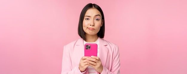 Empresária duvidosa de terno segurando smartphone fazendo careta fazendo expressão de rosto cético em pé sobre fundo rosa