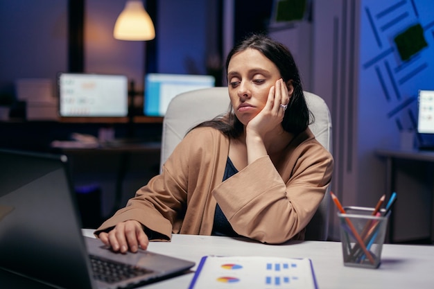 Empresária exausta, olhando para o computador trabalhando para terminar um prazo. Mulher inteligente sentada no local de trabalho durante as primeiras horas da noite, fazendo seu trabalho.