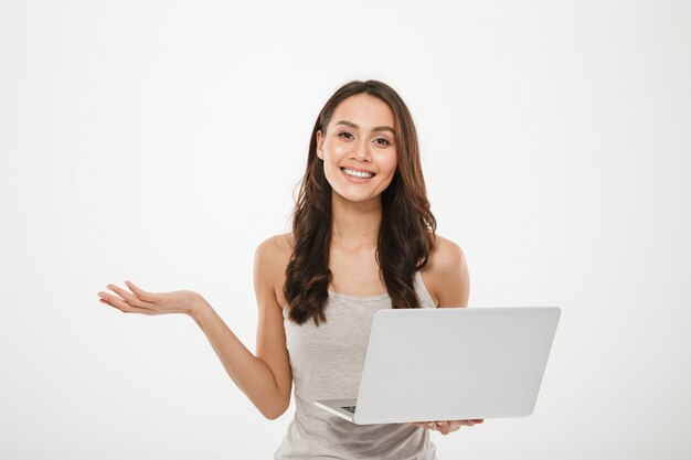 empresária incrível segurando laptop prata e gesticulando com sorriso, sobre parede branca