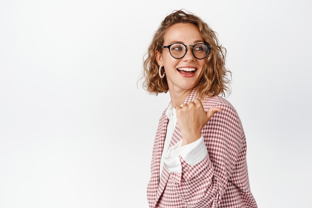 Empresária loira sorridente, gerente de escritório de óculos e terno, apontando e olhando para a direita, atrás do ombro no espaço da cópia, fundo branco.