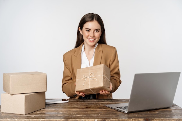 Foto grátis empresária, lojista sentado perto do laptop, segurando a caixa com o pedido do cliente e sorrindo, trabalhando no computador, fundo branco.