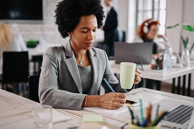 Empresária negra sorridente com uma xícara de café trabalhando em sua mesa e tomando notas em seu caderno Há pessoas no fundo