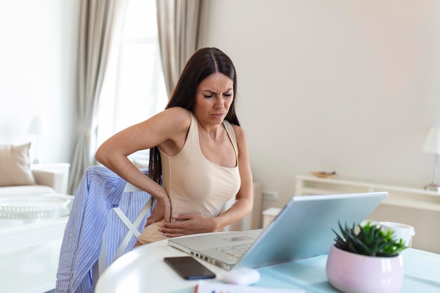 Empresária segurando suas costas enquanto trabalhava no laptop na mesa de escritório em casa jovem esticando sentado sentindo as costas cansadas depois de trabalhar no laptop pequeno interior do escritório em casa