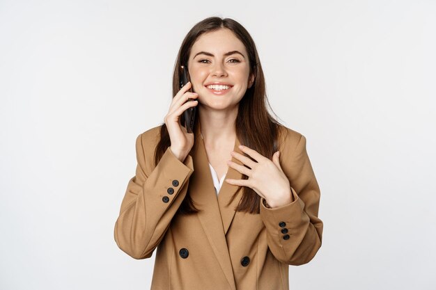 Empresária tendo chamada móvel, conversa no smartphone, conversando com o cliente, de pé sobre fundo branco em terno marrom