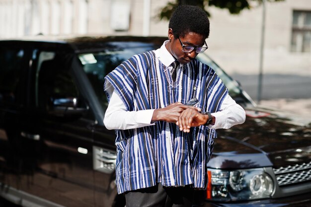 Empresário africano em roupas tradicionais e óculos com celular contra carro preto suv olhando em seus relógios Africanos ricos