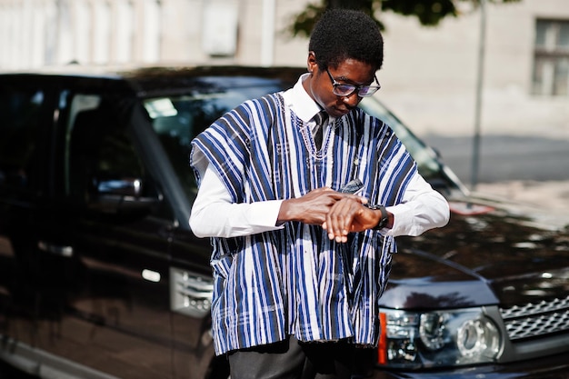 Empresário africano em roupas tradicionais e óculos com celular contra carro preto suv olhando em seus relógios Africanos ricos