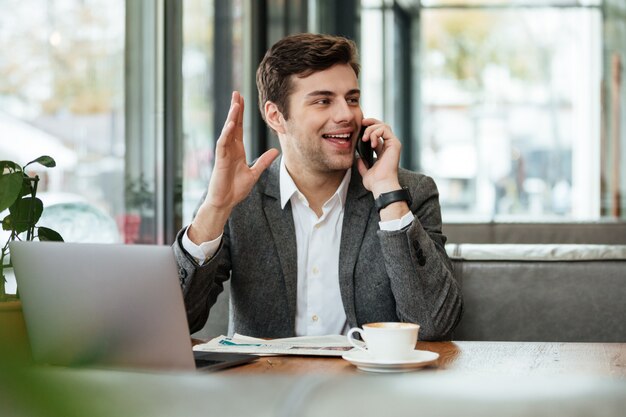 Empresário alegre sentado junto à mesa no café com o computador portátil e falando pelo smartphone enquanto acenando e olhando para longe