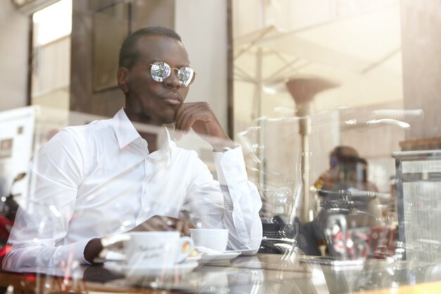 Empresário americano africano moderno sério tomando café no café, sentado à mesa com caneca e olhando através do vidro da janela do lado de fora, segurando a mão no queixo com expressão pensativa e pensativa