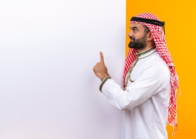 Empresário árabe com roupa tradicional com outdoor em branco apontando o dedo para cima e sorrindo em pé sobre a parede laranja