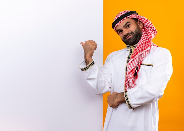 Foto grátis empresário árabe com roupa tradicional em pé perto de um outdoor em branco apontando com o dedo para ele com um sorriso confiante no rosto sobre a parede laranja