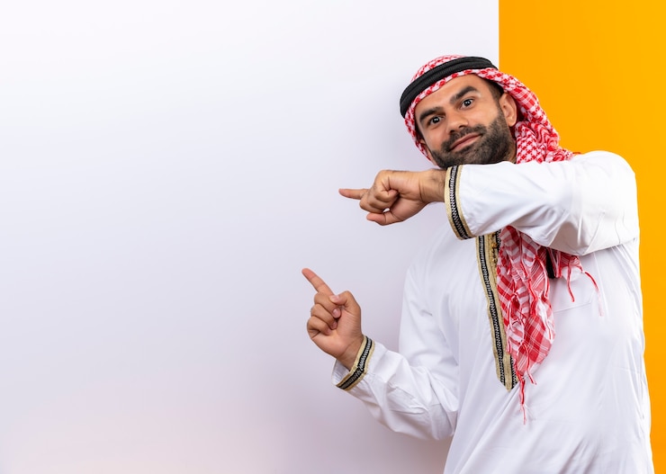 Foto grátis empresário árabe com roupa tradicional em pé perto de um outdoor em branco apontando com os dedos para ele com um sorriso confiante no rosto sobre a parede laranja