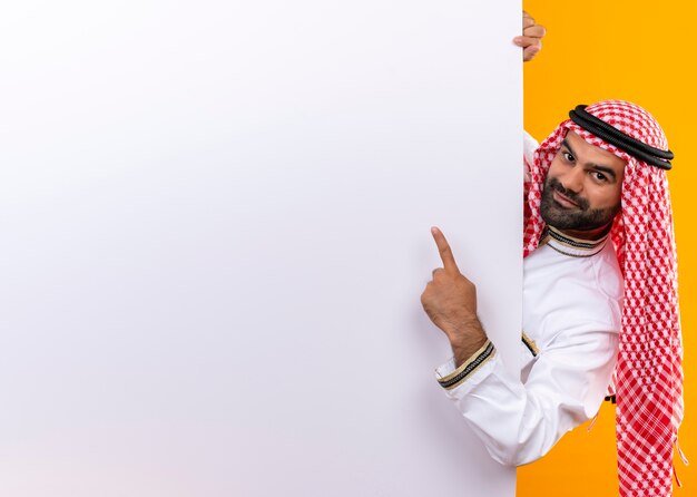 Empresário árabe com roupa tradicional espiando outdoor em branco apontando com o dedo e sorrindo em pé sobre a parede laranja