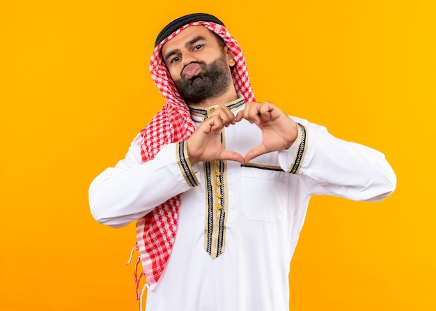 Empresário árabe com roupa tradicional fazendo gestos de coração com os dedos sobre o peito tentando dar um beijo em pé sobre a parede laranja