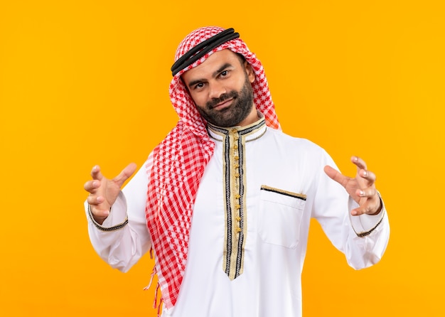 Empresário árabe com roupa tradicional, mostrando um gesto de tamanho grande com as mãos sorrindo com confiança, símbolo de medida em pé sobre a parede laranja