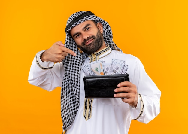 Empresário árabe com roupa tradicional segurando carteira com dinheiro sorrindo confiante apontando com o dedo para o dinheiro em pé sobre a parede laranja