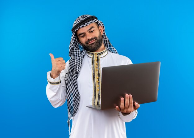 Empresário árabe com roupa tradicional segurando laptop olhando para a tela de seu laptop apontando para trás com um sorriso no rosto em pé sobre a parede azul