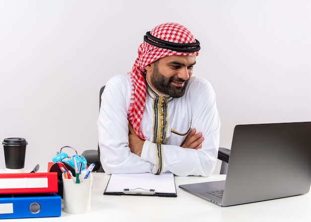 Foto grátis empresário árabe com roupa tradicional sentado à mesa com um laptop olhando para a tela, sorrindo, trabalhando no escritório