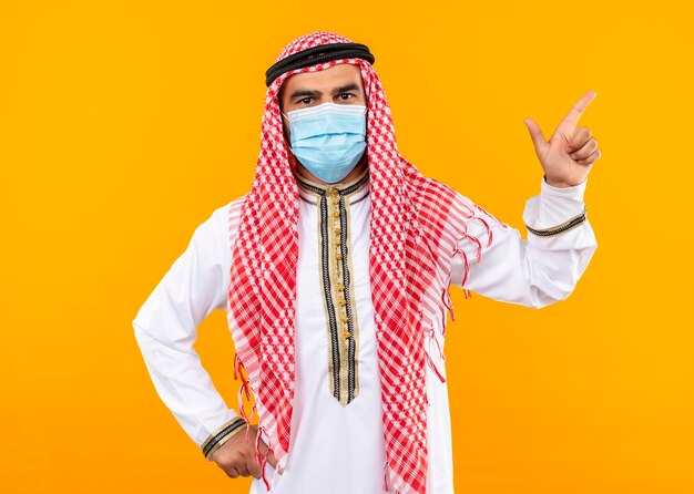 Empresário árabe em trajes tradicionais e máscara protetora facial com expressão confiante apontando para o lado com o dedo indicador em pé sobre a parede laranja