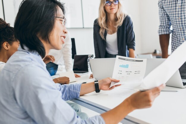 Empresário asiático sorridente analisando o infográfico em seu escritório. Retrato interno de jovens especialistas em TI com um desenvolvedor chinês de óculos