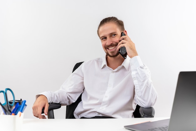 Empresário bonito em camisa branca falando no celular sorrindo sentado à mesa em escritório sobre fundo branco