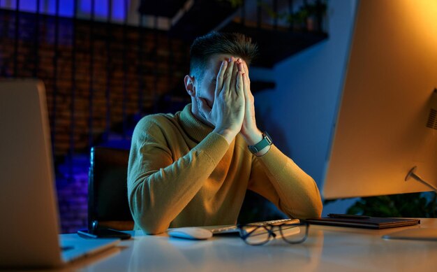 Empresário cansado de depressão, sentado no escritório tarde da noite após horas, trabalhando no computador e laptop, cobrindo o rosto com as mãos. freelancer, trabalhando em casa à noite.