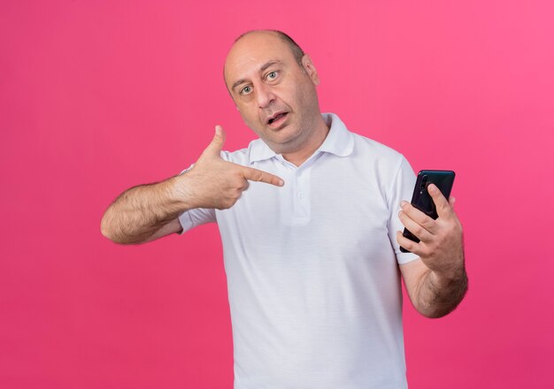 empresário casual maduro impressionado segurando e apontando para o celular isolado em rosa