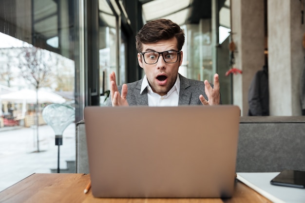 Empresário chocado em óculos, sentado junto à mesa no café, olhando para o computador portátil