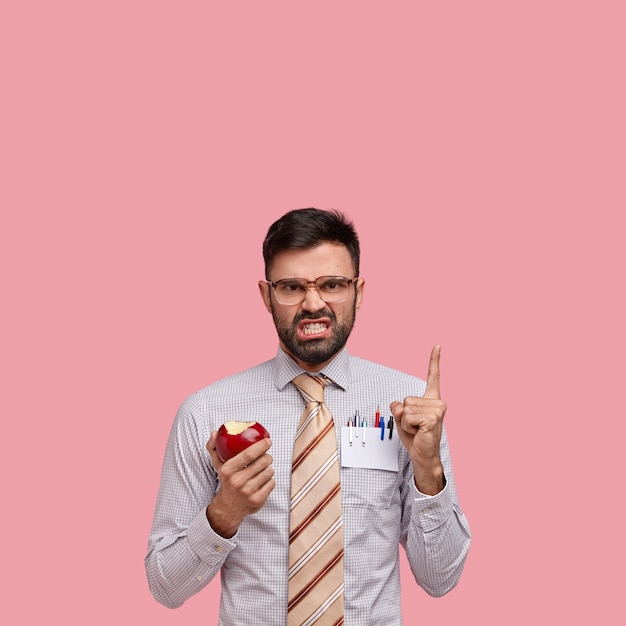 Foto grátis empresário com a barba por fazer irritado aponta para o espaço em branco acima, franze a testa em descontentamento, usa óculos grandes, come maçã vermelha