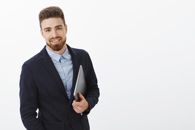Empresário com olhos azuis e barba em pé, confiante em um terno formal, segurando o laptop na mão, olhando satisfeito e seguro, sendo ambicioso e bem-sucedido