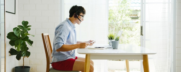 Empresário da Ásia vestido com camiseta e shorts usa laptop para falar com colegas em videochamada enquanto trabalha em casa na sala de estar. Auto-isolamento, distanciamento social, quarentena para prevenção do vírus corona.