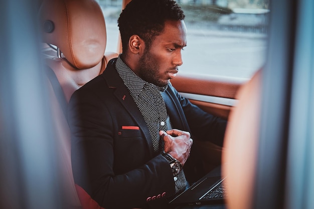 Empresário de etnicidade afro elegante pensativo está sentado no carro como passageiro enquanto trabalha em seu laptop.