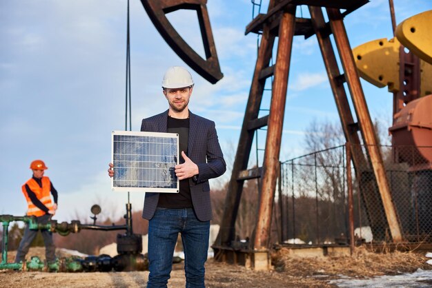 Empresário de pé em um campo petrolífero segurando mini módulo solar ao lado de uma plataforma de petróleo