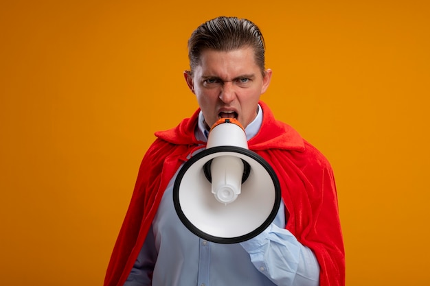 Empresário de super-herói zangado com capa vermelha gritando para o megafone com expressão agressiva em pé sobre a parede laranja