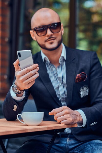 Empresário elegante atraente em óculos de sol está sentado no café do lado de fora enquanto conversa no celular.