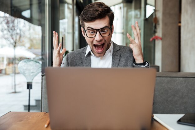 Empresário gritando chocado em óculos, sentado junto à mesa no café e se alegrar enquanto olha para o computador portátil