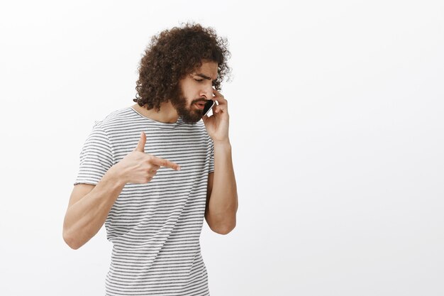 empresário hispânico bonito e mandão com barba e penteado afro, virando à direita enquanto fala no smartphone e gesticulando com uma arma de dedo