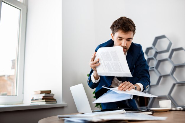 Empresário jovem distraído remexendo nos papéis, fundo do escritório.