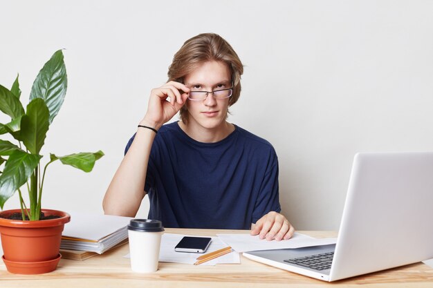 Empresário masculino assegurado olha seriamente através dos vidros, cercado com o laptop moderno