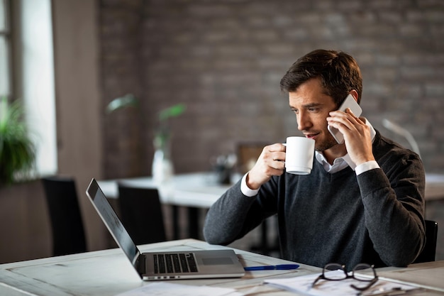 Empresário masculino falando no celular enquanto bebe café e navega na net em um computador no escritório