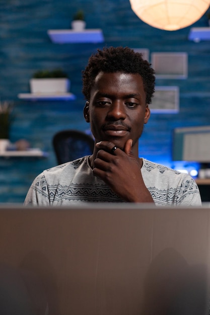 Empresário procurando informações para lição financeira usando computador portátil trabalhando remotamente de casa. Adulto afro-americano sentado no sofá lendo curso para projeto de comunicação