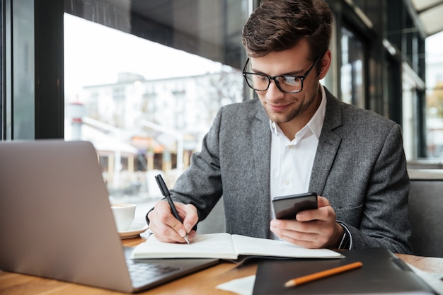 Empresário sorridente em óculos, sentado junto à mesa no café com o computador portátil enquanto estiver usando o smartphone e escrevendo algo