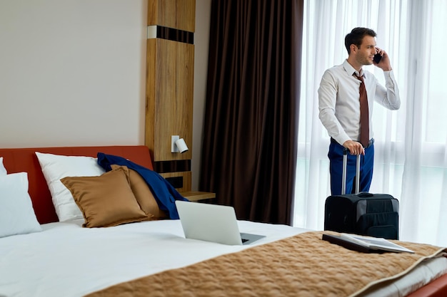Empresário sorridente se comunicando no celular em pé com uma mala de viagem no quarto de hotel