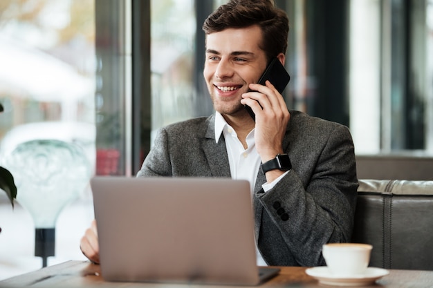 Empresário sorridente, sentado junto à mesa no café com o computador portátil enquanto fala pelo smartphone