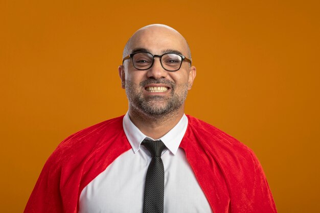 Foto grátis empresário super-herói com capa vermelha e óculos, olhando para a frente, sorrindo amplamente feliz e positivo em pé sobre a parede laranja