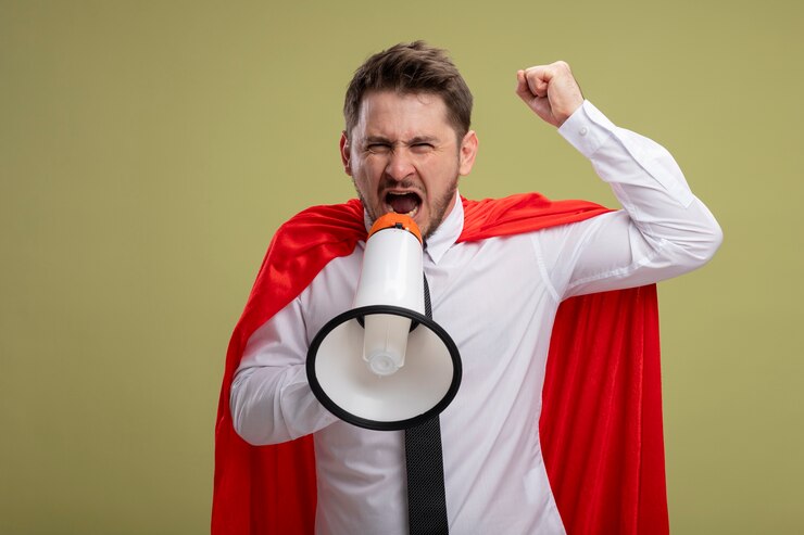 Foto grátis empresário super-herói com capa vermelha gritando para o megafone com expressão agressiva levantando o punho em pé sobre a parede verde