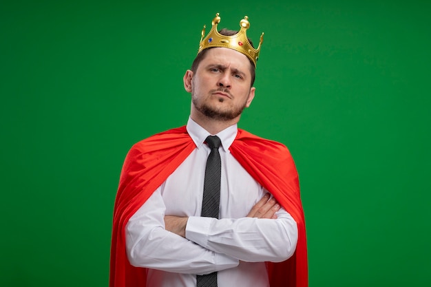 Foto grátis empresário super-herói com capa vermelha usando coroa, olhando para a câmera com expressão séria e confiante com os braços cruzados no peito em pé sobre fundo verde