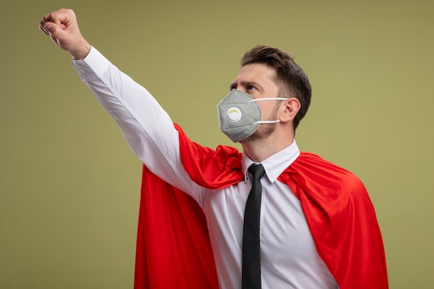 Foto grátis empresário super-herói com máscara facial protetora e capa vermelha, fazendo um gesto vencedor, parecendo confiante, pronto para ajudar em pé sobre a parede verde