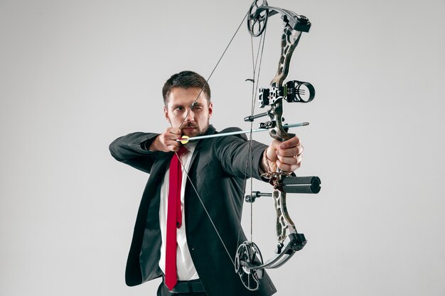 Empresário visando o alvo com arco e flecha, isolado em fundo cinza.