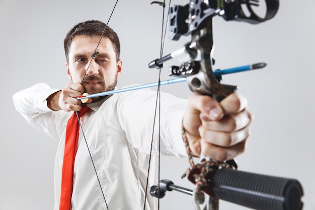 Empresário visando o alvo com arco e flecha, isolado em fundo cinza.